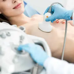 Lekarz od piersi wykonuje badanie USG. Dłoń w niebieskiej rękawiczce trzyma głowicę aparatu, przykładając ją do klatki piersiowej pacjentki.