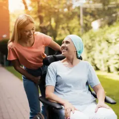 Uśmiechnięta kobieta w chuście na głowie siedzi na wózku inwalidzkim, a młoda kobieta pcha go. Nadzieja, że czy rak 4 stopnia jest wyleczalny, dodaje im sił.