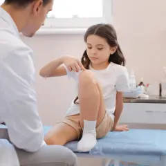 Dziewczynka wskazuje lekarzowi ból kolana. Trwa diagnostyka, czy to zwykły uraz, czy może ból kolana u dziecka związany z nowotworem.