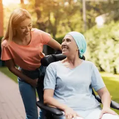 Uśmiechnięta kobieta w chuście na głowie, siedząca na wózku inwalidzkim, cieszy się spacerem z młodszą kobietą. To piękny obraz nadziei i remisji choroby.