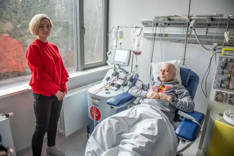 Kobieta w czerwonej bluzce stoi obok pacjenta podłączonego do aparatury medycznej. To ważny moment w procesie przeszczepu szpiku.