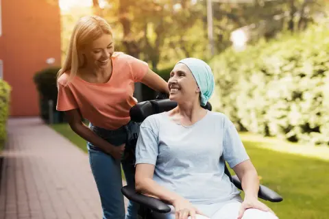 Uśmiechnięta kobieta w chuście na głowie siedzi na wózku inwalidzkim, a młoda kobieta pcha go. Nadzieja, że czy rak 4 stopnia jest wyleczalny, dodaje im sił.