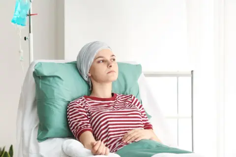 Młoda kobieta w chuście na głowie leży w szpitalnym łóżku, czekając na kolejną chemię. Czerwona w białe paski koszulka kontrastuje z zielonymi poduszkami.