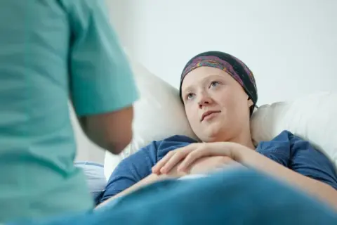 Młoda kobieta w chustce na głowie leży w łóżku szpitalnym, rozmawiając z pielęgniarką. Długie zwolnienie po operacji oszczędzającej jest nieuniknione.