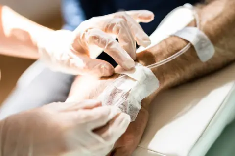 Ręce w rękawiczkach zakładają opatrunek na wenflon. Pacjent przechodzi chemioterapię, która bywa trudna, ale każdy cykl przybliża do zdrowia.