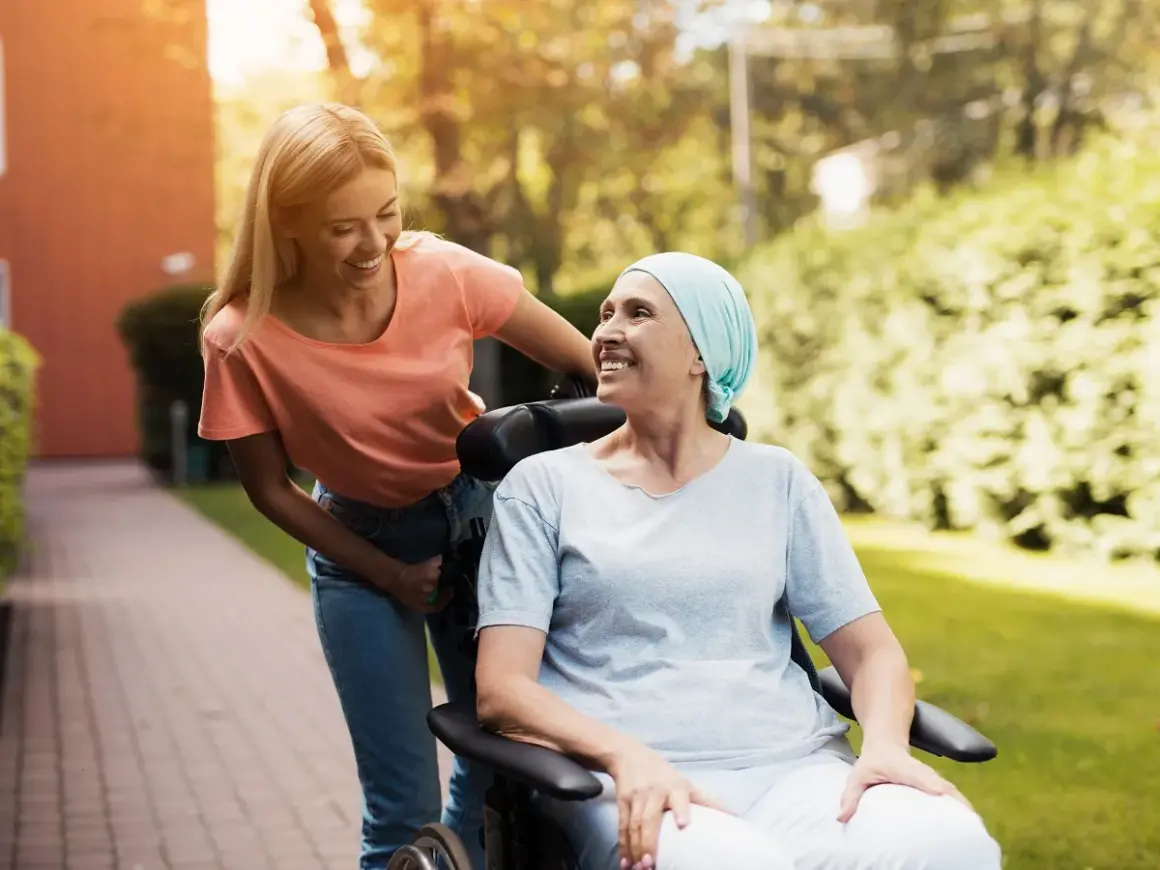 Uśmiechnięta kobieta w chuście na głowie siedzi na wózku inwalidzkim, a młoda kobieta pcha go. Nadzieja, że czy rak 4 stopnia jest wyleczalny, dodaje im sił.