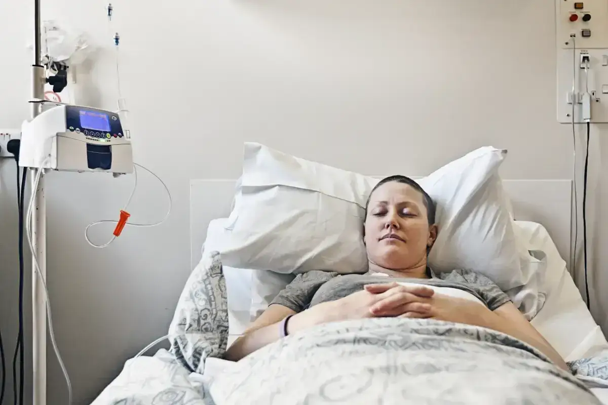 Kobieta z ogoloną głową leży w łóżku szpitalnym, podłączona do kroplówki. Jej oczy są zamknięte, dłonie złożone. To obraz walki z rakiem piersi w 4 stadium, gdzie rokowania są trudne.